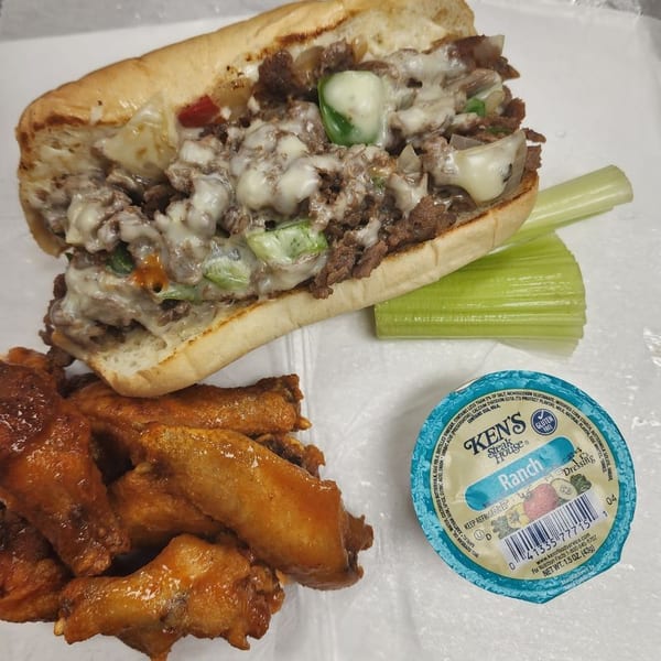 Cheesesteak sandwich with melted cheese, green peppers, and onions, served with chicken wings, celery, and ranch dressing.