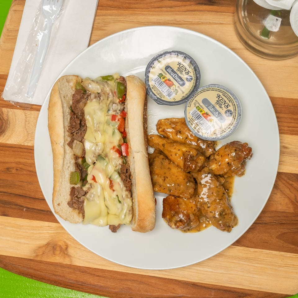Philly cheesesteak with melted cheese, peppers, and onions, served with glazed chicken wings and dressings.