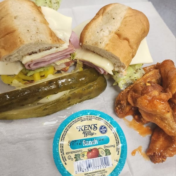 A sub sandwich, a pickle spear, buffalo wings, and a Ken's Ranch dressing cup on white paper.